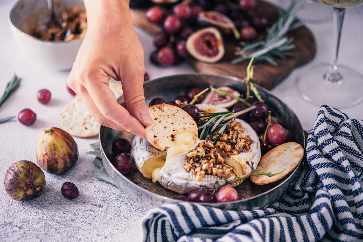 A woman dips a cracker into a wheel of brie garnished with herbs and fruit
