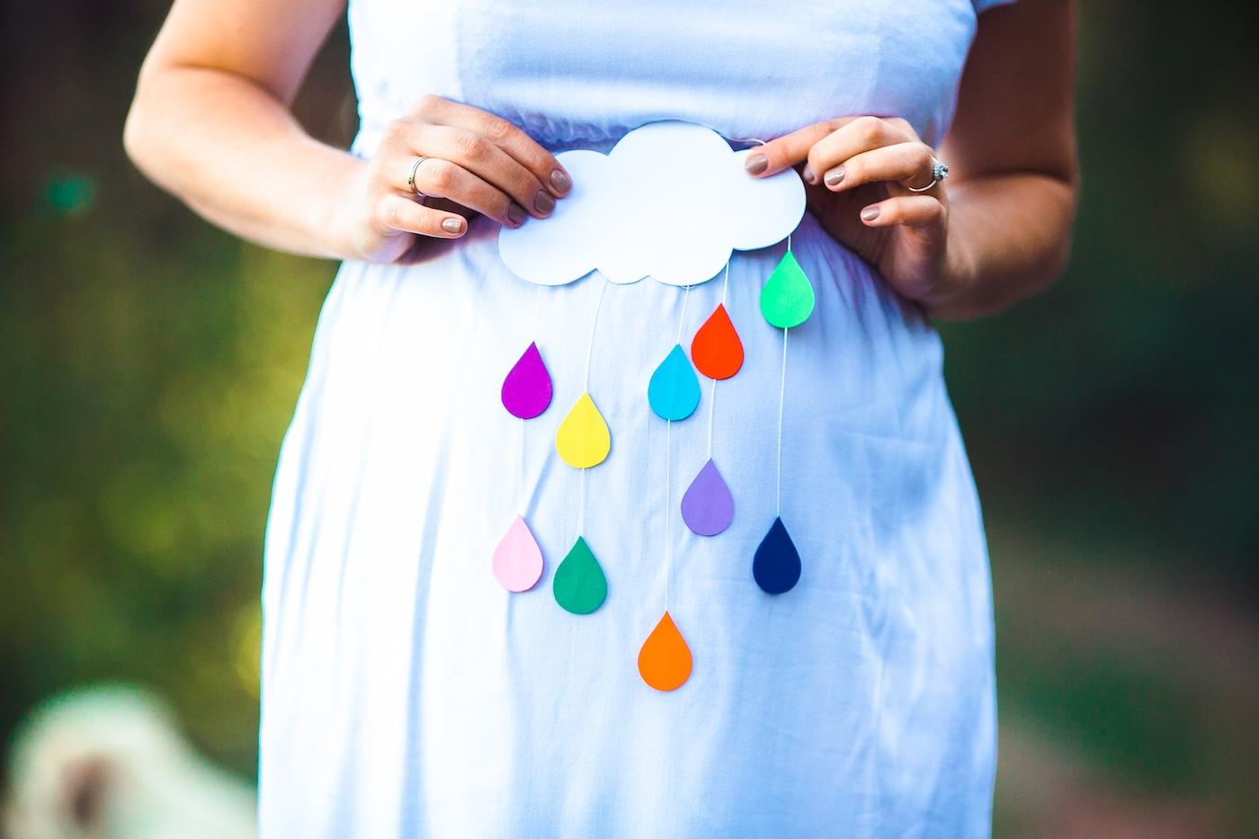 A pregnant woman holds up a paper cloud with rainbow colored raindrops over her baby bump to announce a rainbow baby