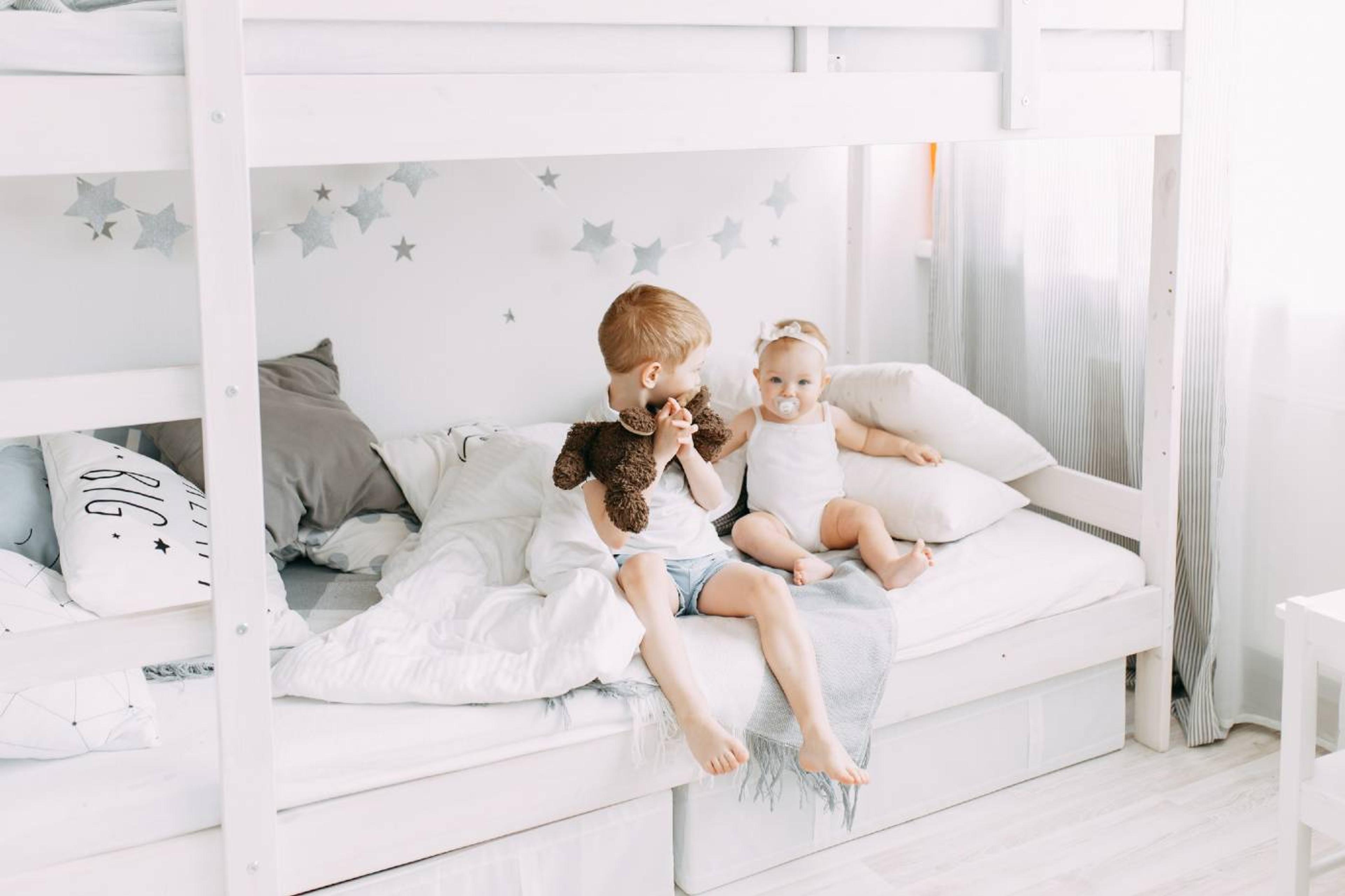A young boy and his toddler sister sit on the bottom of a bunk bed.