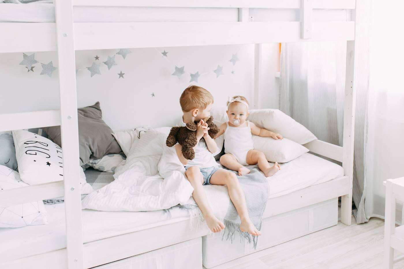 A young boy and his toddler sister sit on the bottom of a bunk bed.