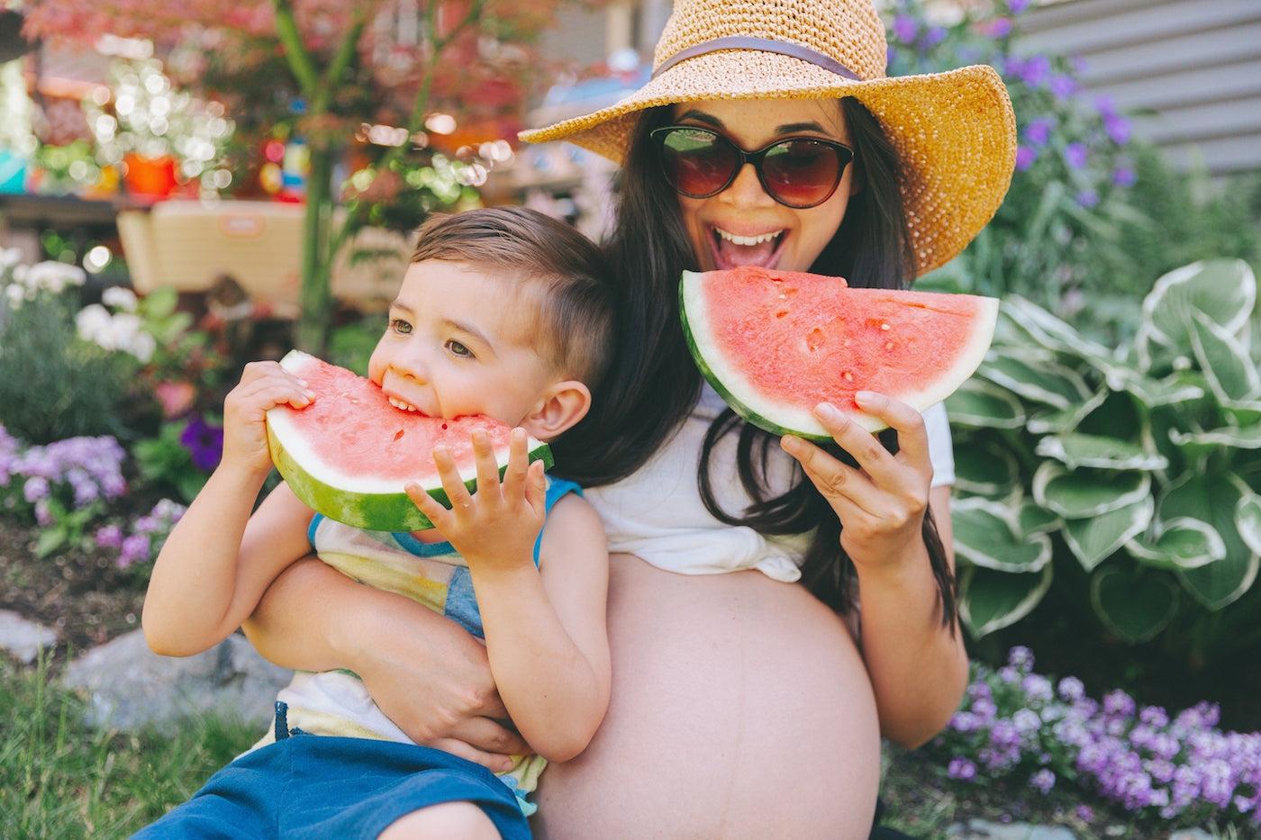 A pregnant mom and her toddler son eat watermelon together outside