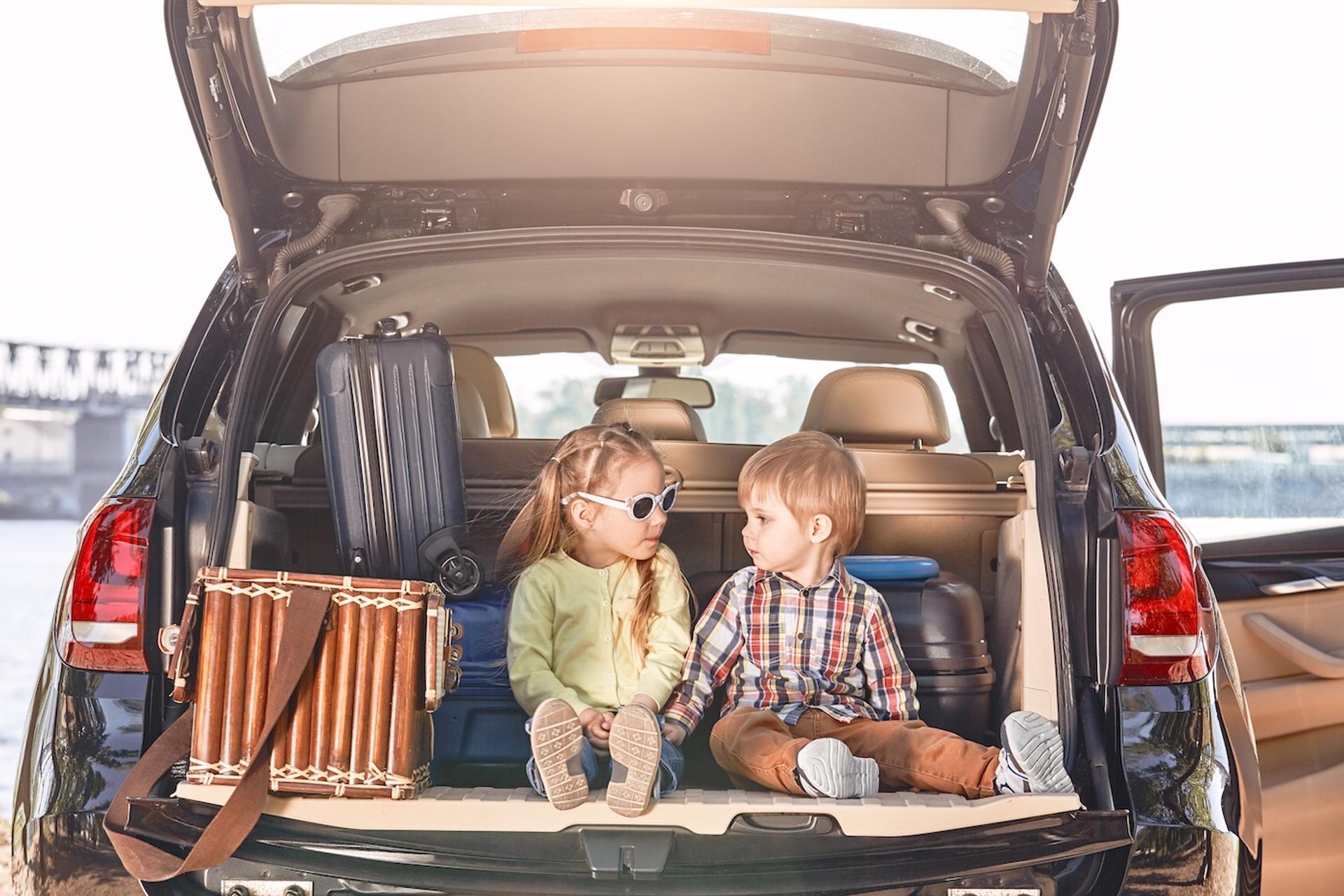 Two little kids sit in the back of their car at a rest stop during a family road trip