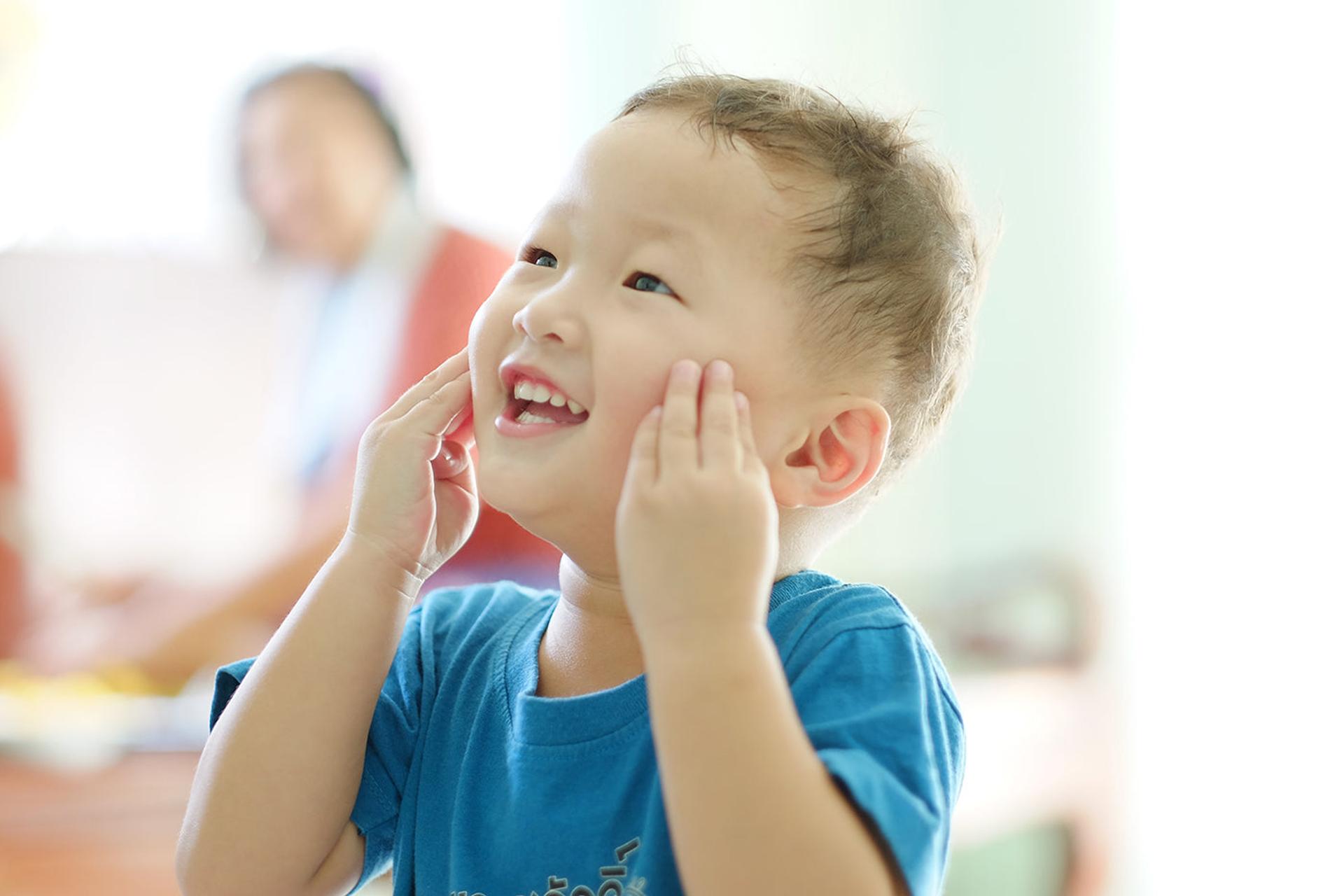 A young Korean boy smiles and touches his face