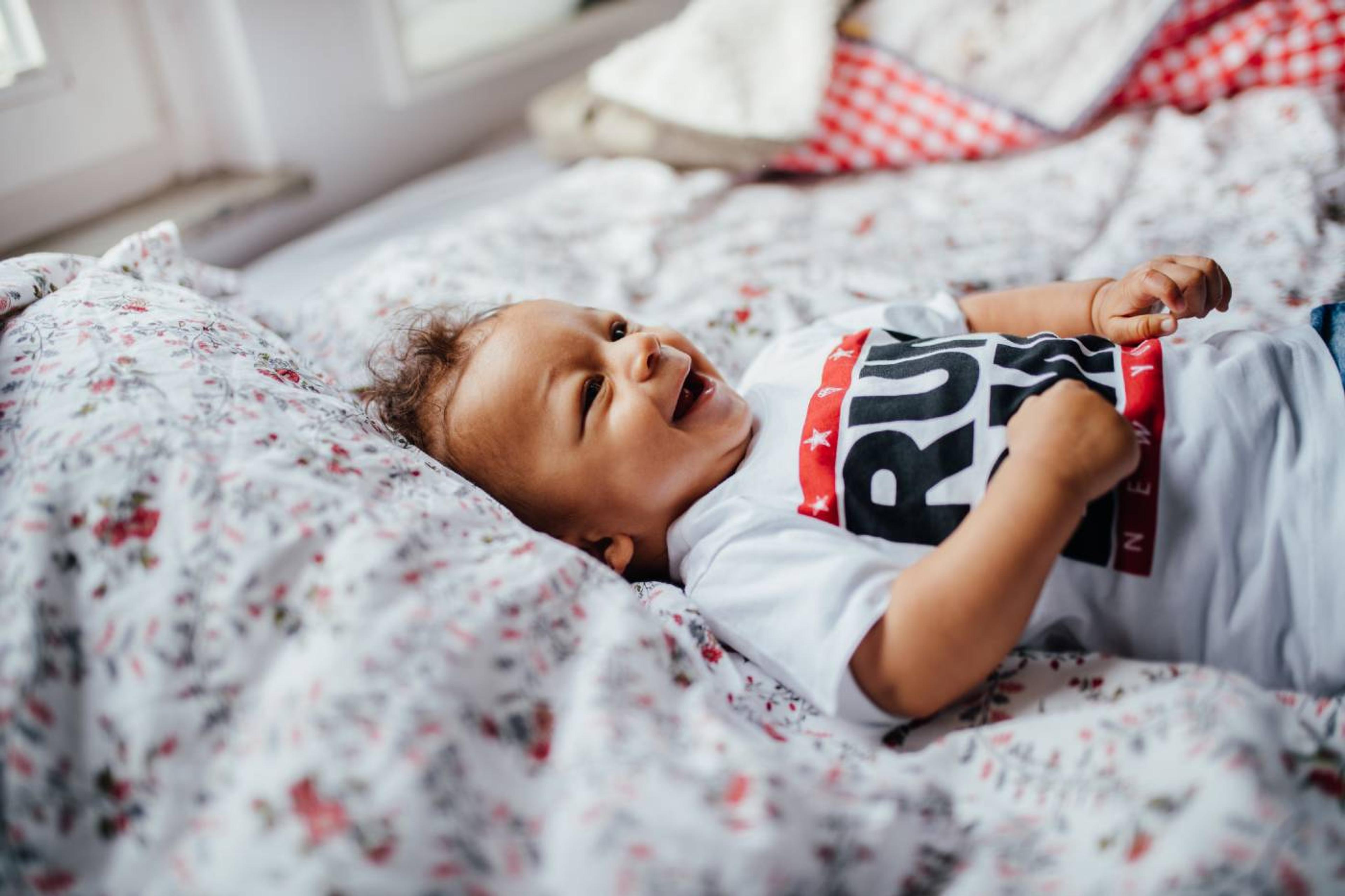 A baby boy wearing a "Run DMC" bodysuit lays on a bed and smiles
