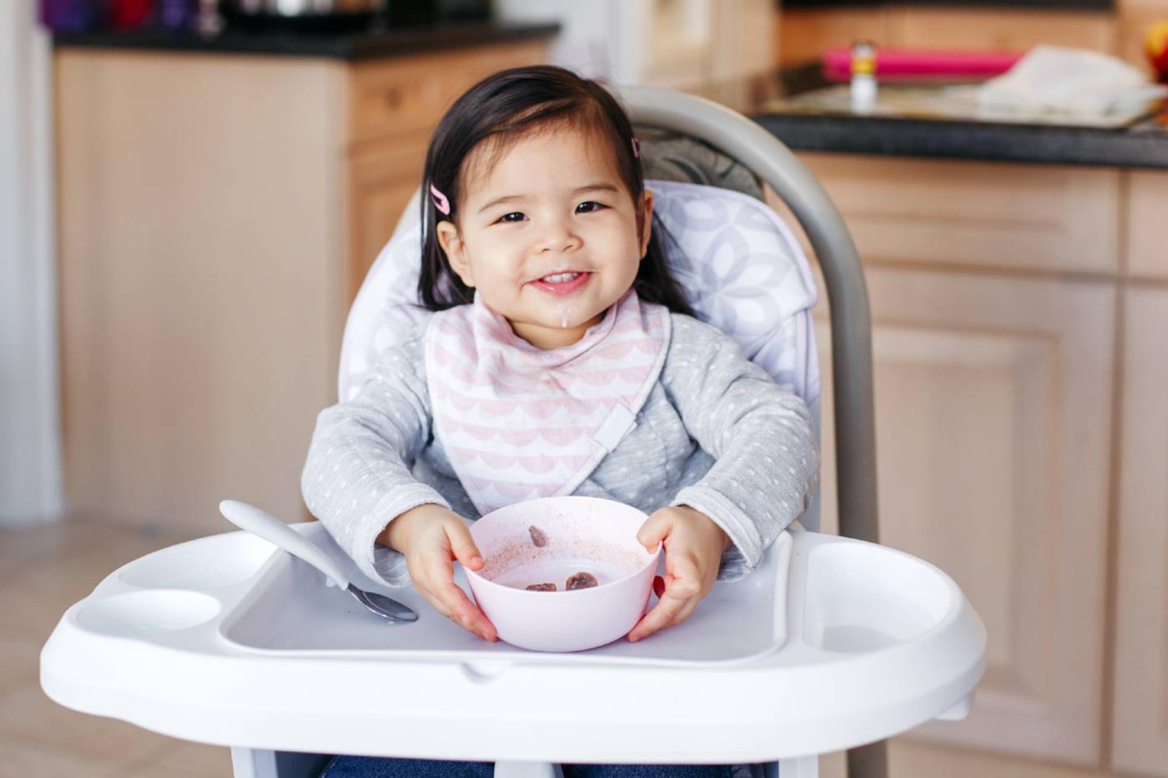 Toddler girl smiles as she sits in a highchair and holds a bowl of food