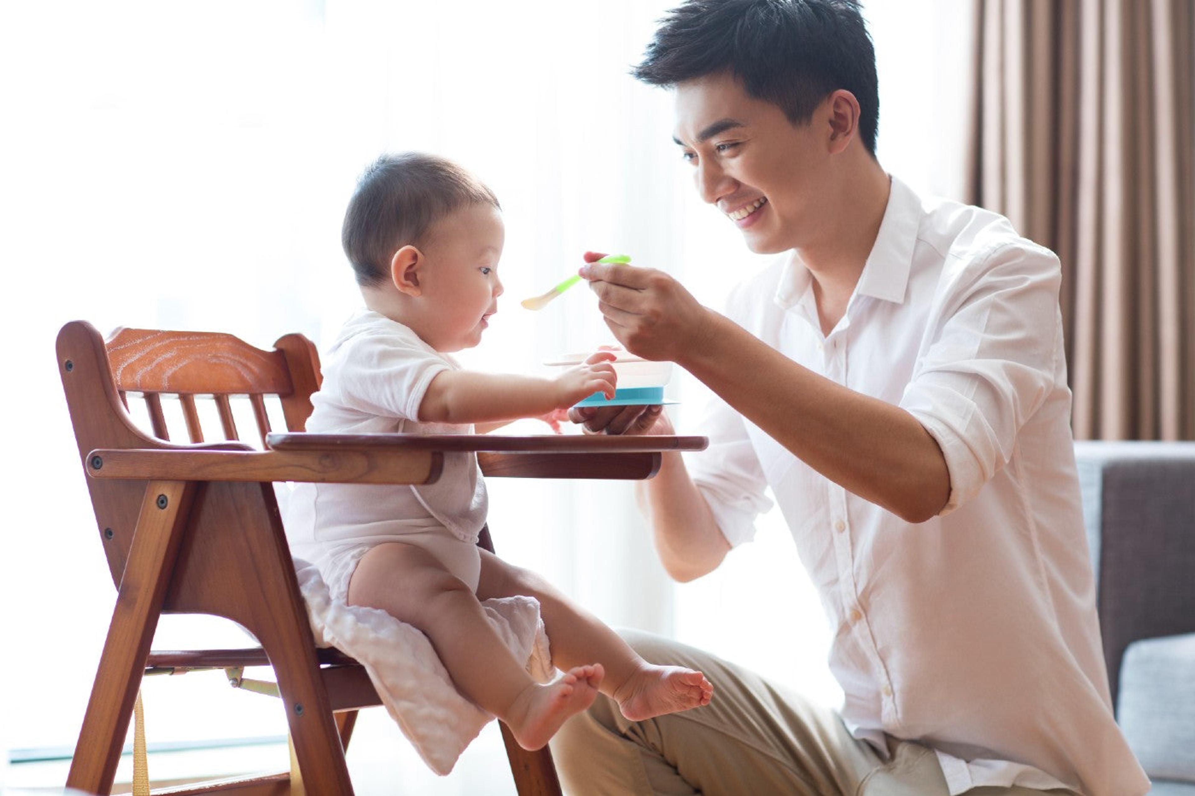 A father spoon-feeds a baby in a highchair