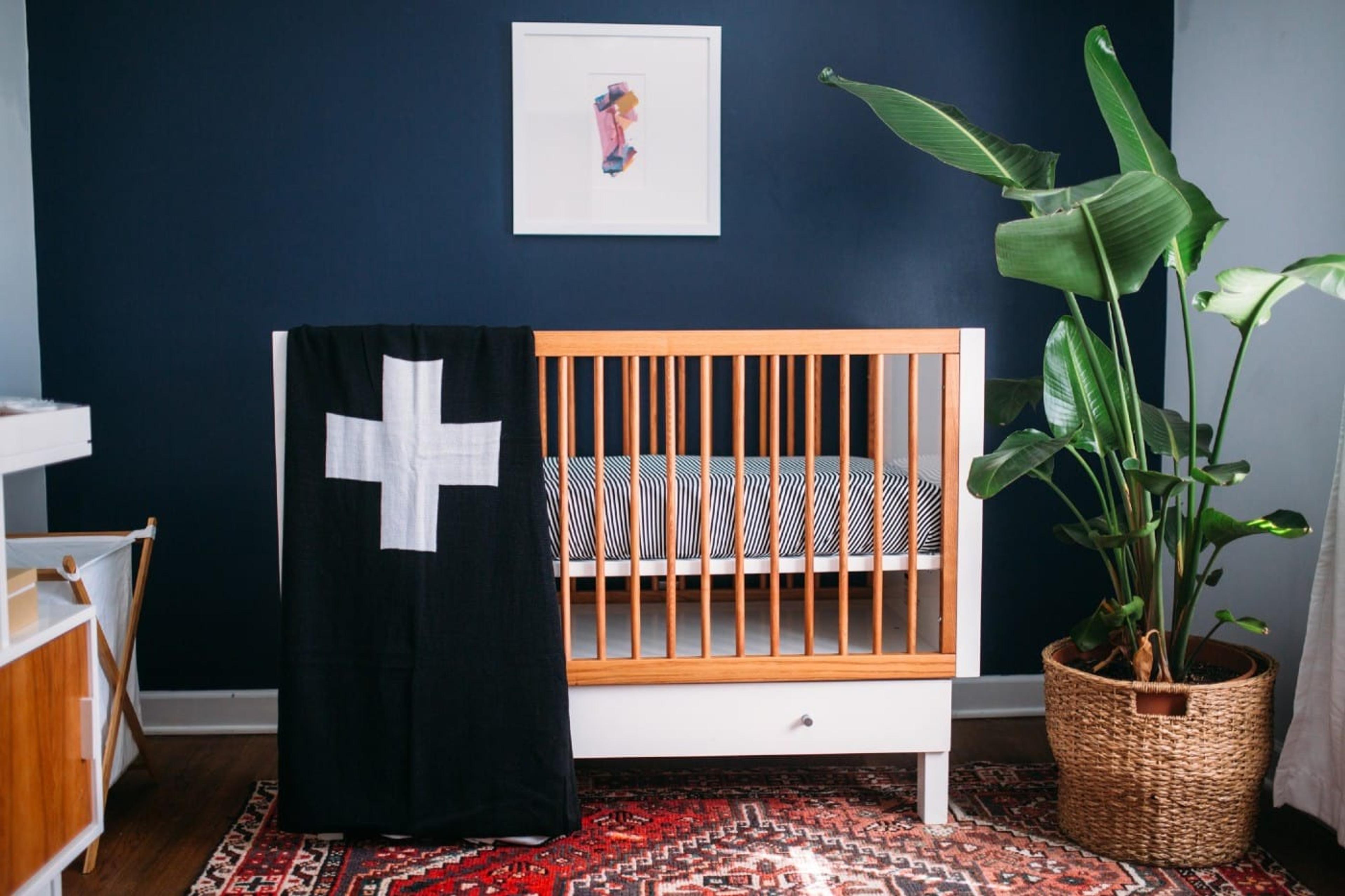 A white and walnut crib with a blanket draped over it against a dark blue wall next to a houseplant.