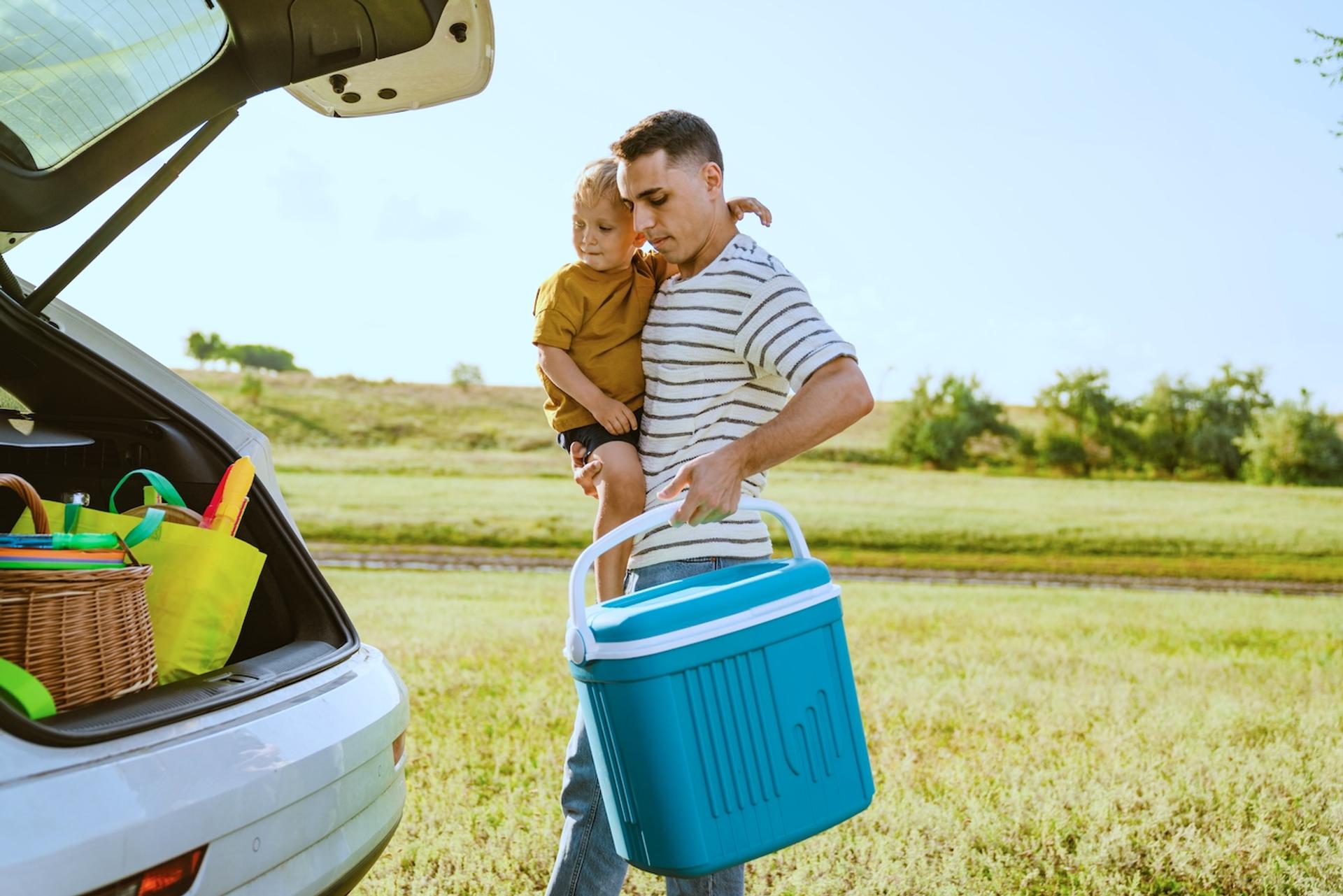 A dad holding a toddler puts a cooler full of travel snacks in the back of his car