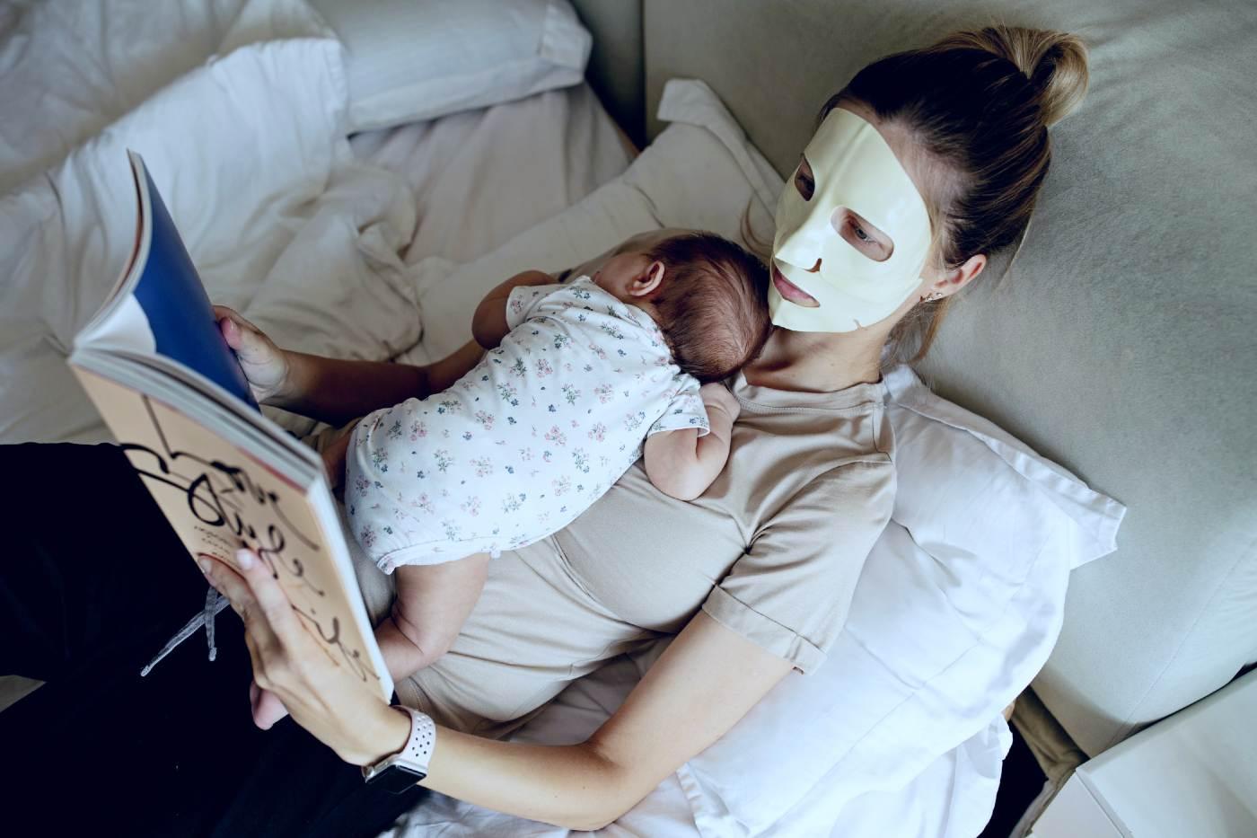 postpartum self-care: a new mom wearing a face mask and reading while a baby sleeps on her chest