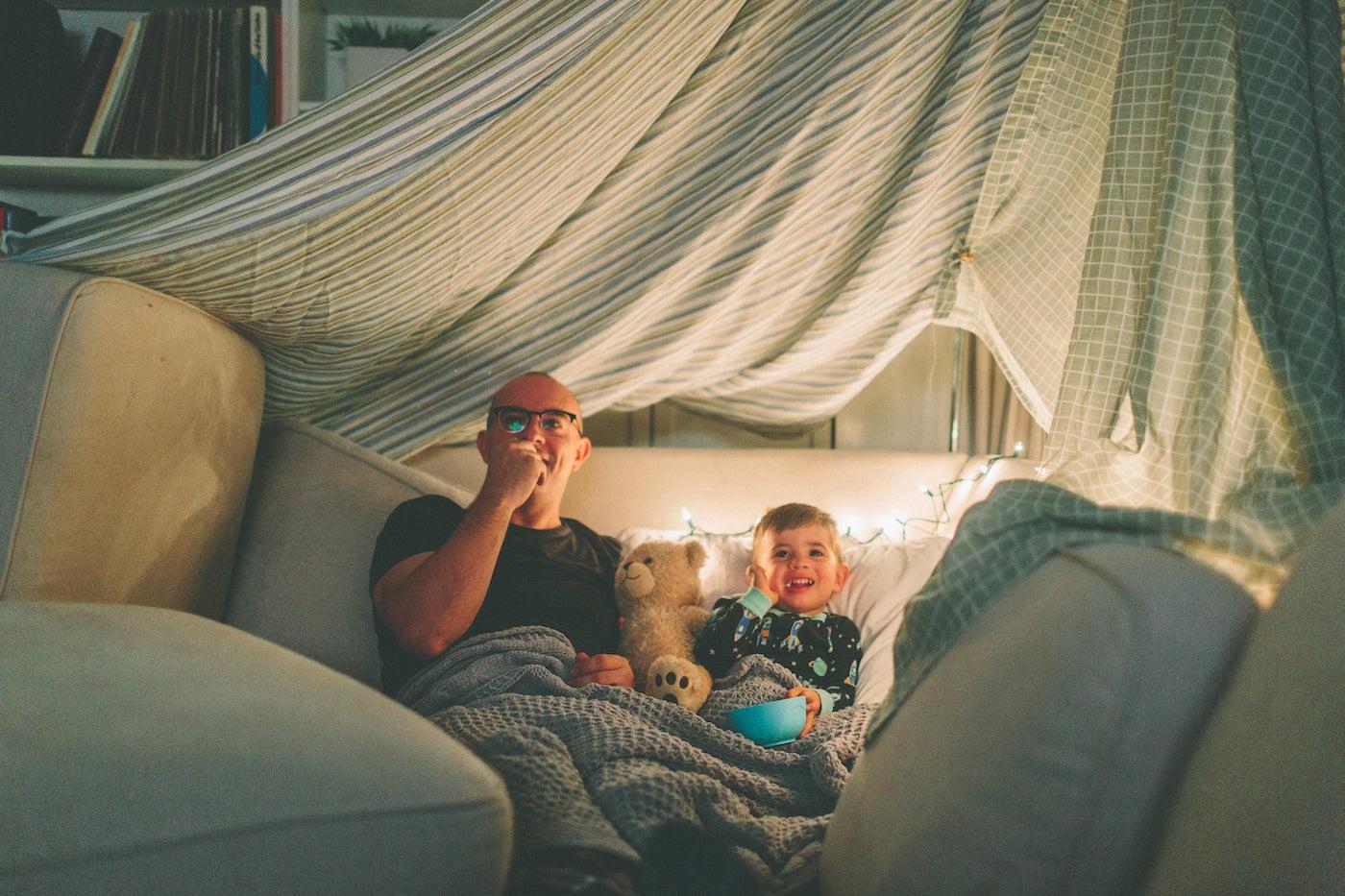 A father and son watch a movie in a homemade fort during a family staycation