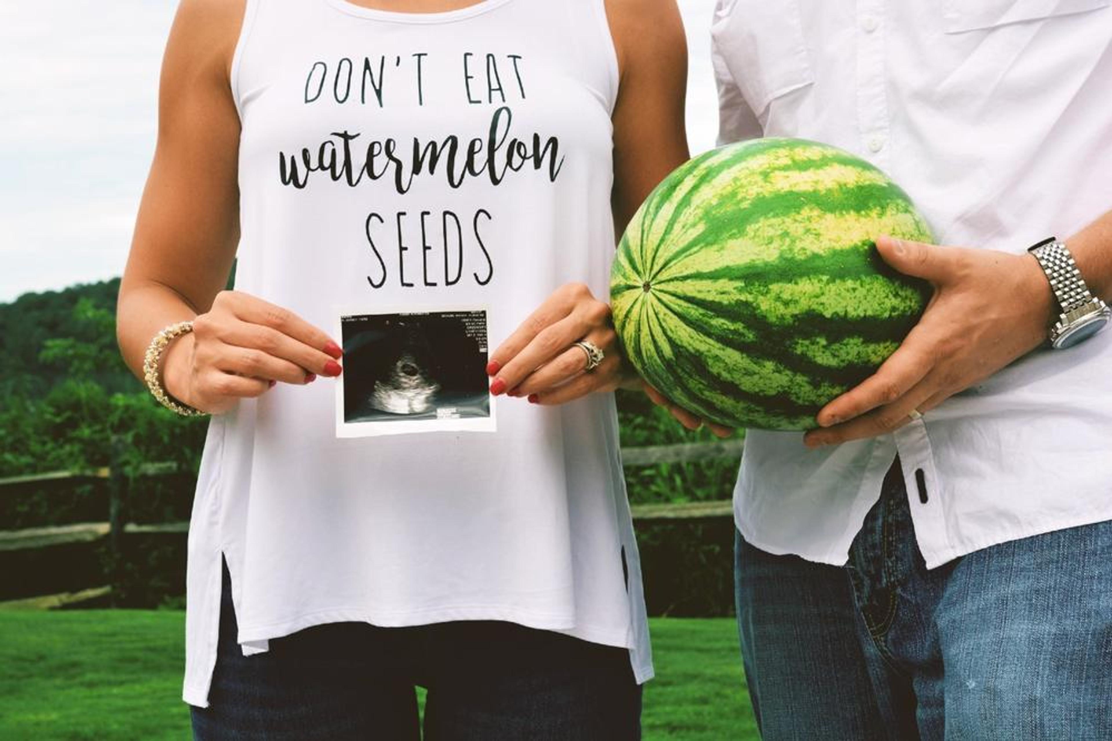 Woman holding ultrasound wearing shirt that says "don't eat watermelon seeds" standing next to a man holding a watermelon