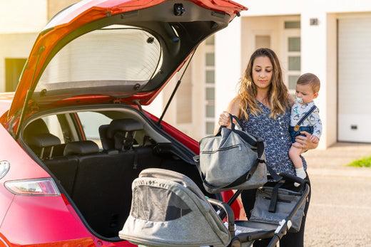 A woman holding a baby organizes baby gear in her car trunk