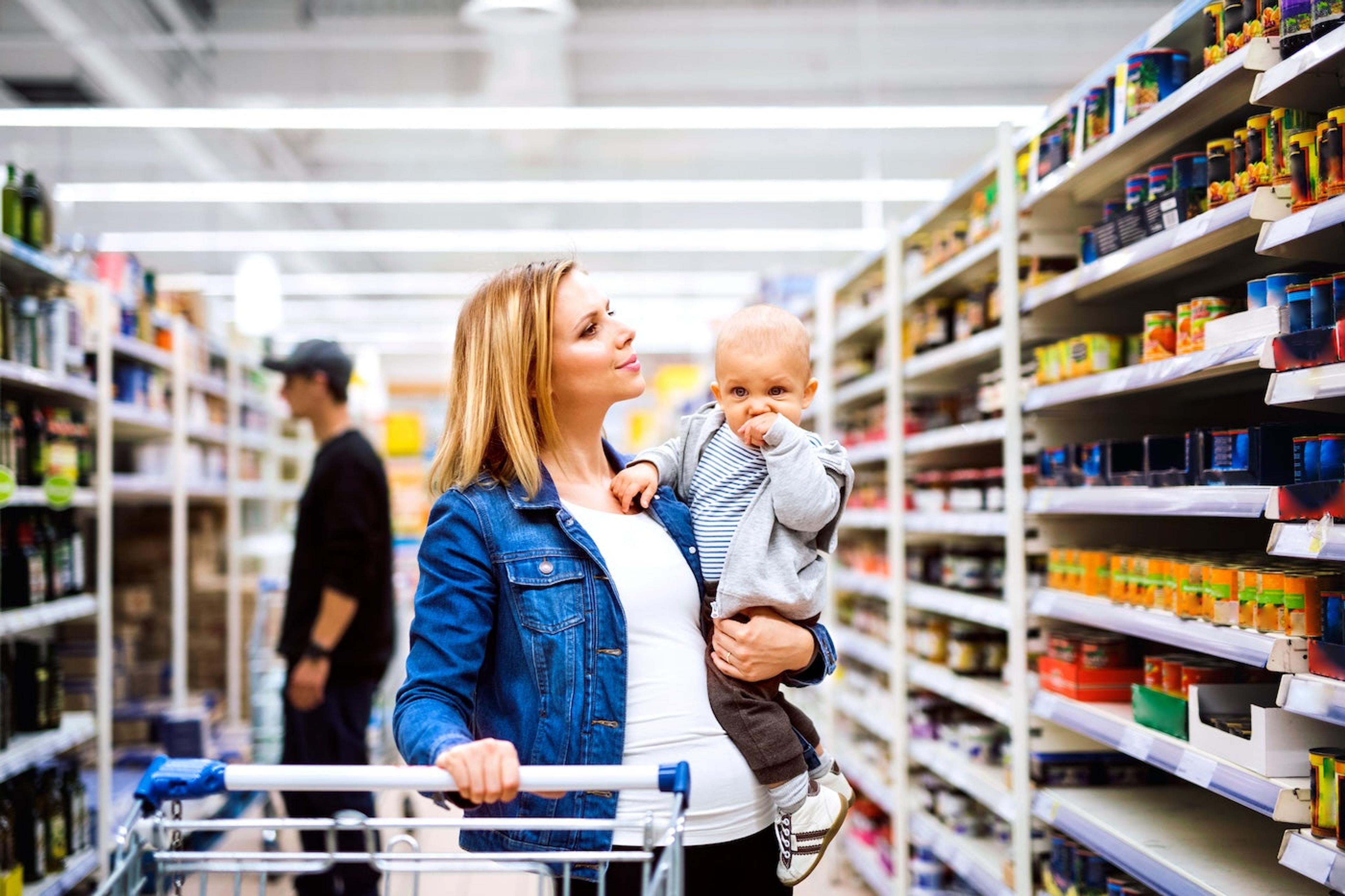A mom examines packaged foods on a grocery store shelf as she holds her young toddler