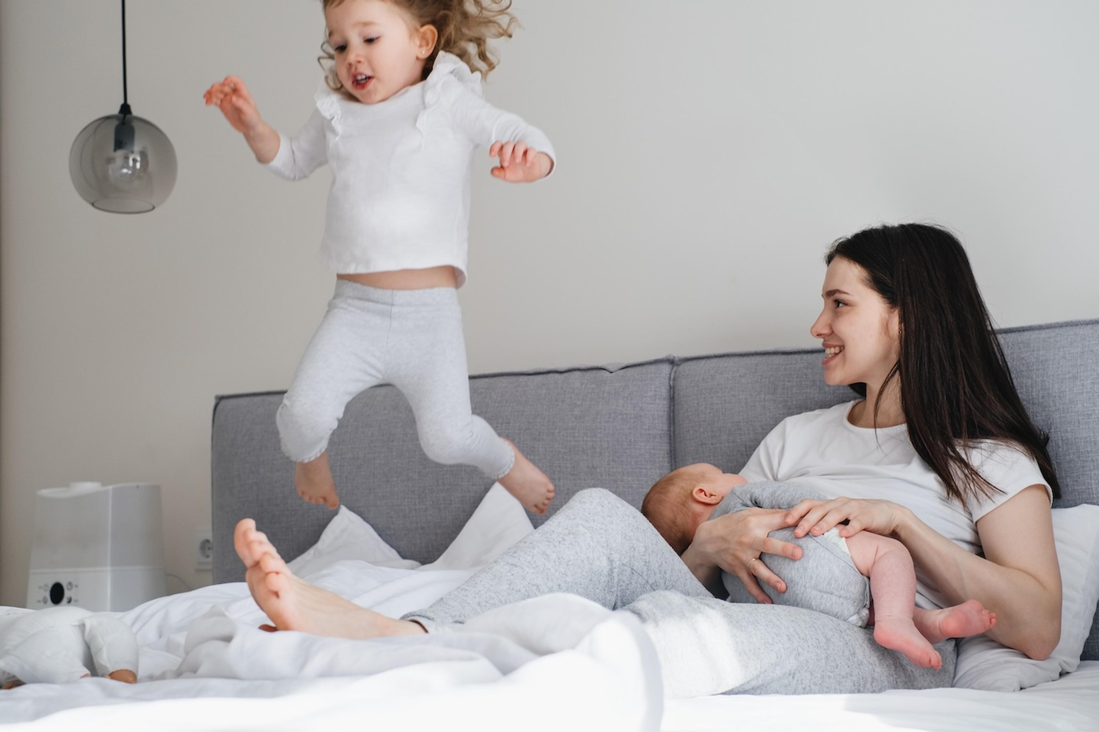 A mother nurses her baby while her toddler jumps on the bed