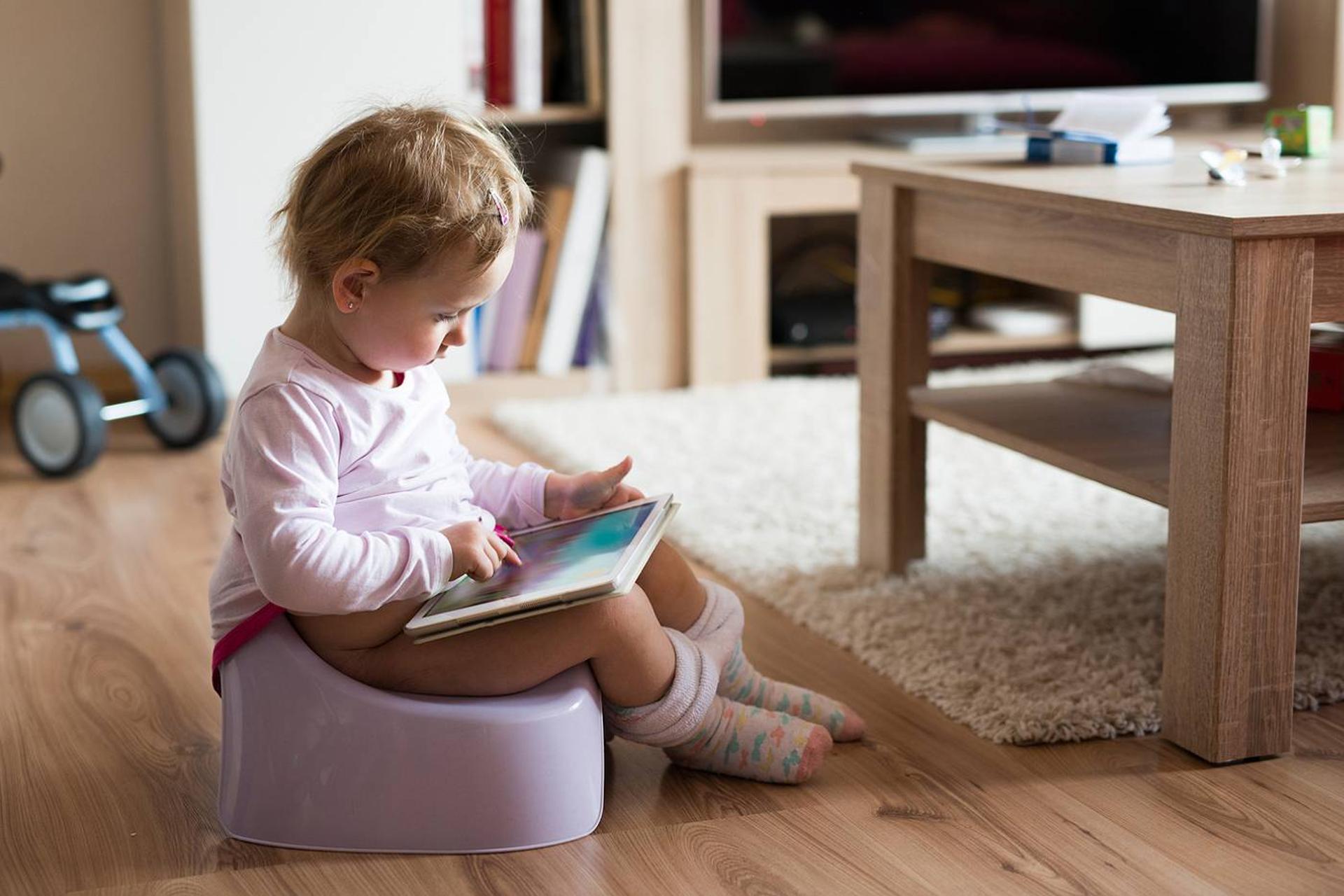 A toddler sits on a small toilet with a book during potty training