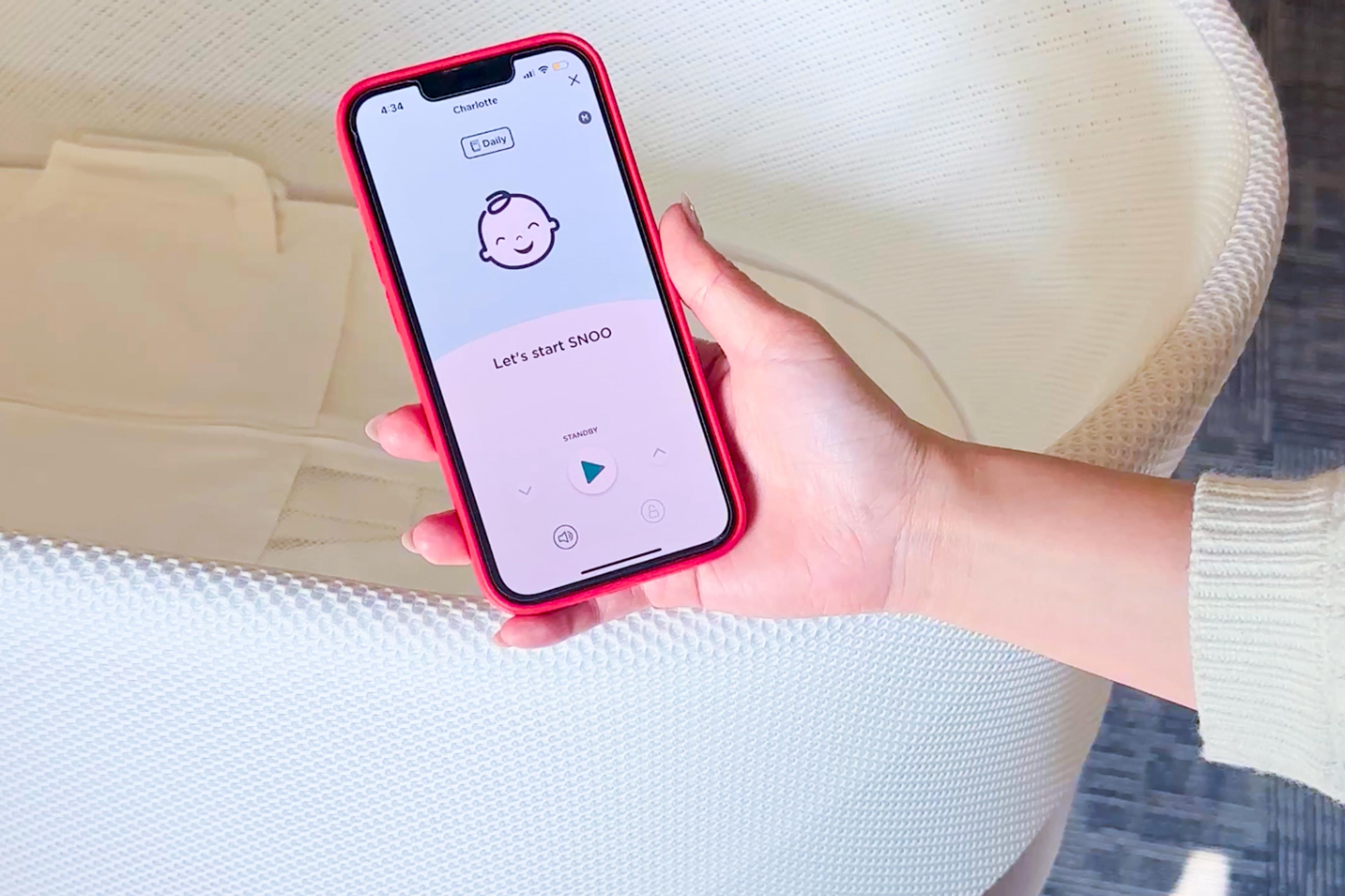 A woman holding a mobile phone with the Happiest Baby app open, controlling the SNOO bassinet