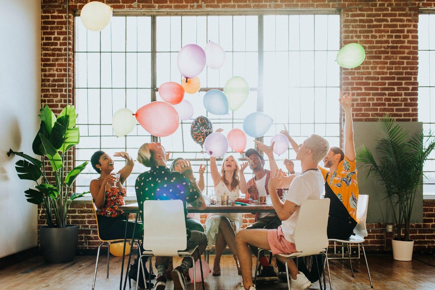 A group of coworkers celebrate an office baby shower around a conference table with balloons in an industrial-loft-style workspace
