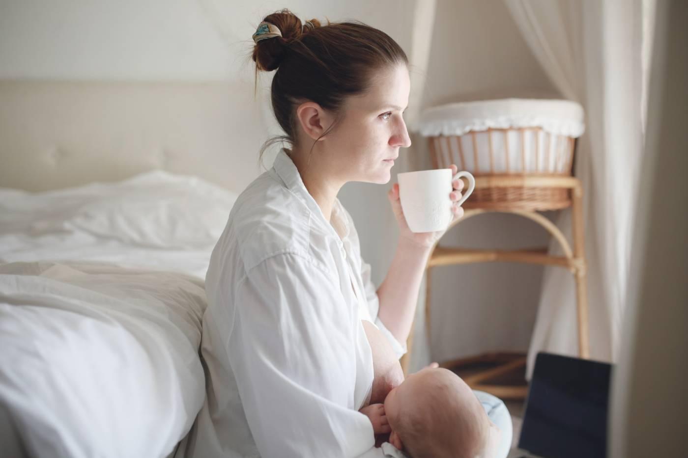Tired mom, holding a coffee mug while nursing a newborn