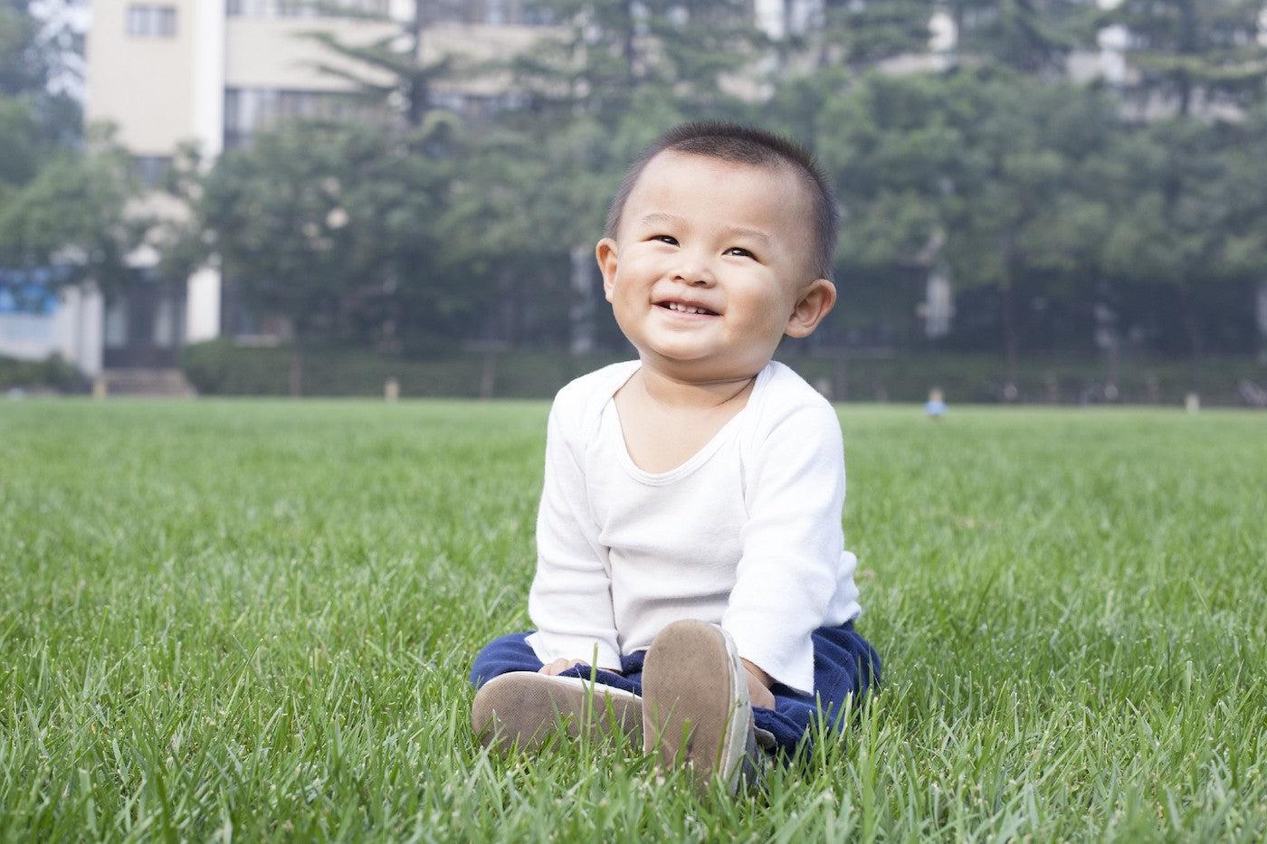 A 17-month-old baby boy sits in the grass and smiles