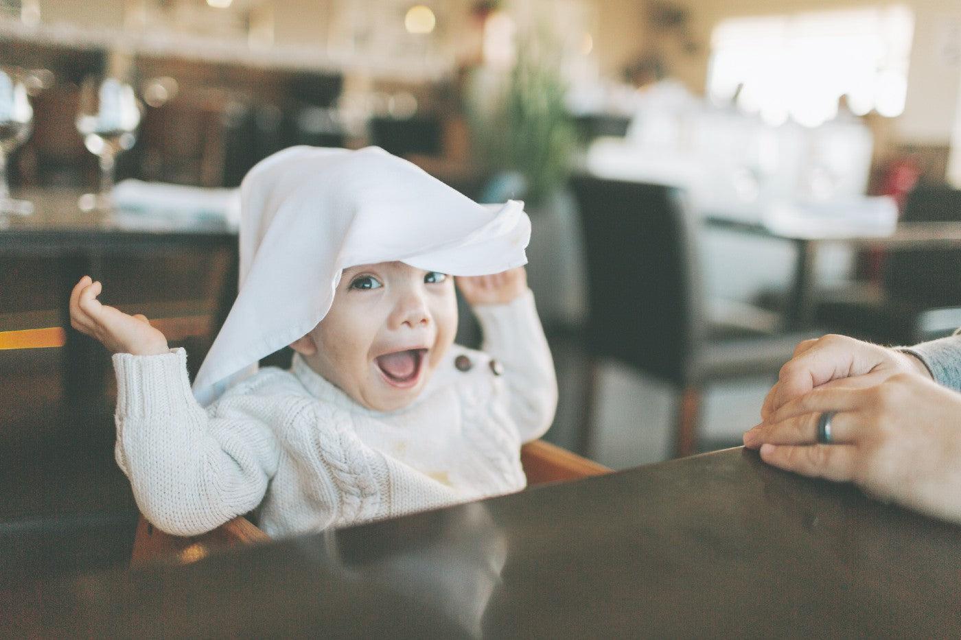 A baby sits in a restaurant highchair with a cloth napkin on his head