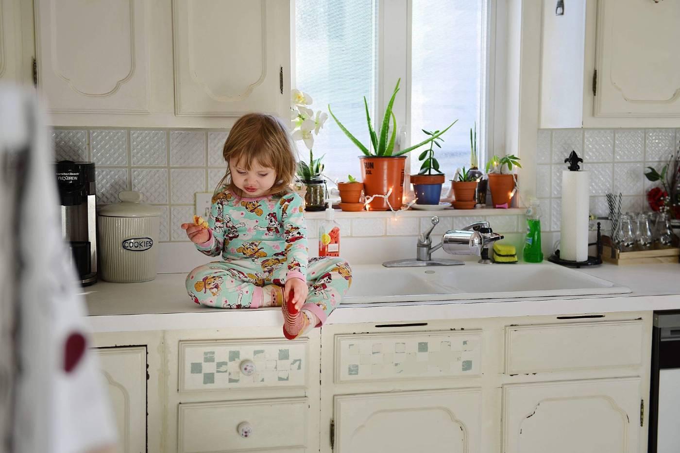 A toddler girl sits on the kitchen counter and eats a snack in her PJs