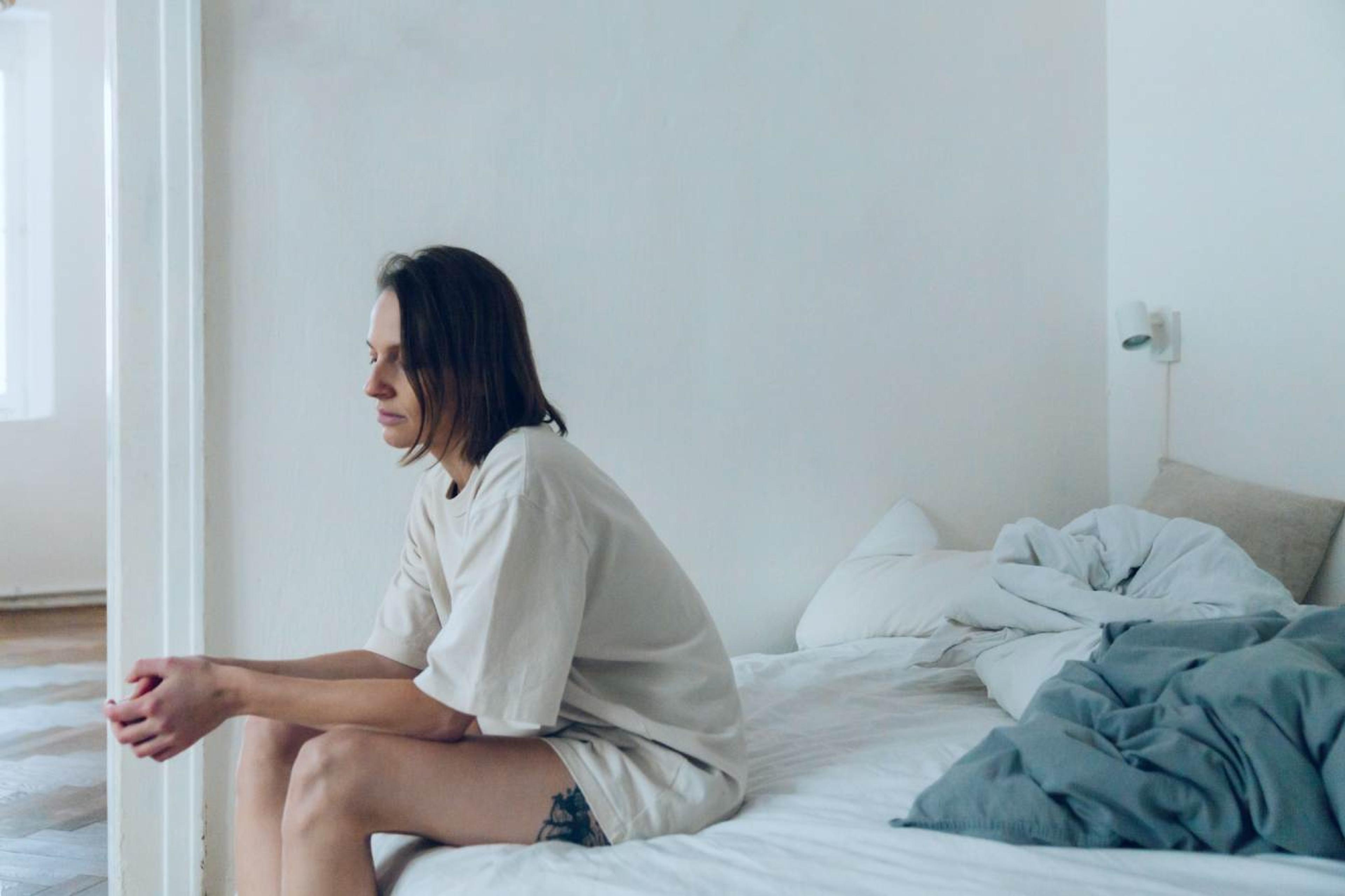 Sad-looking woman sitting on the edge of her bed, looking off into the distance.