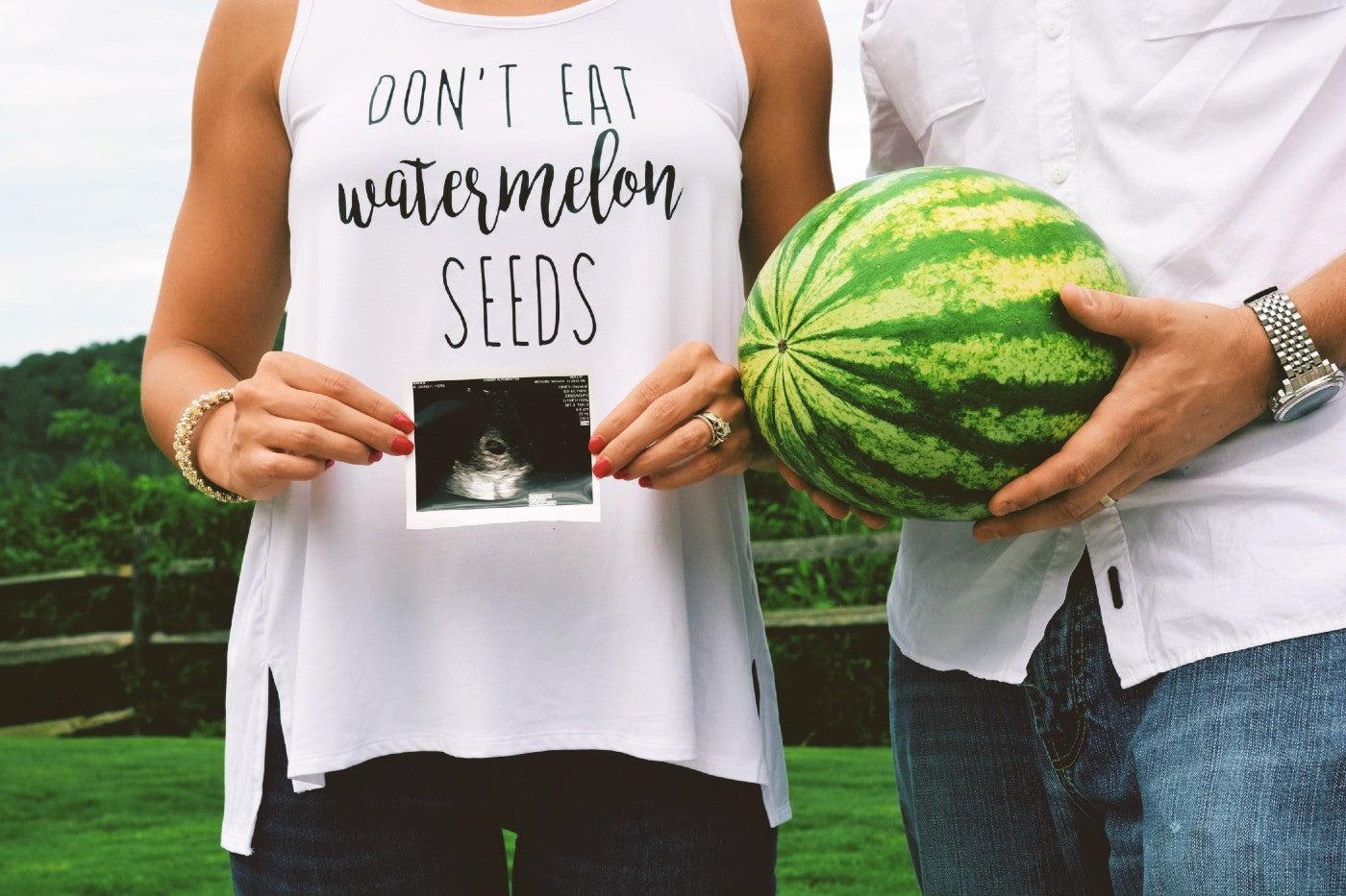 Woman holding ultrasound wearing shirt that says "don't eat watermelon seeds" standing next to a man holding a watermelon