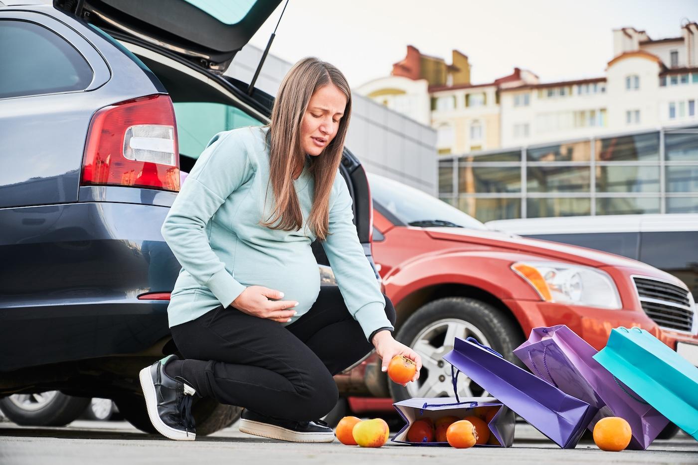 A pregnant woman bends down to pick up fruit that she dropped in a parking lot.