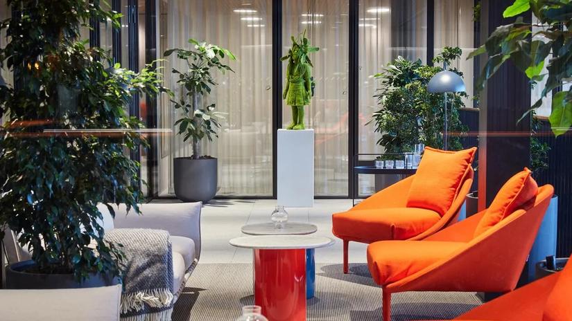 Modern lounge area with vibrant orange chairs, a small round table, and lush green plants, featuring a green statue on a pedestal in the background.