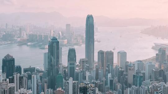 Aerial view of Hong Kong's skyline with tall skyscrapers and Victoria Harbour in the background under a hazy sky.