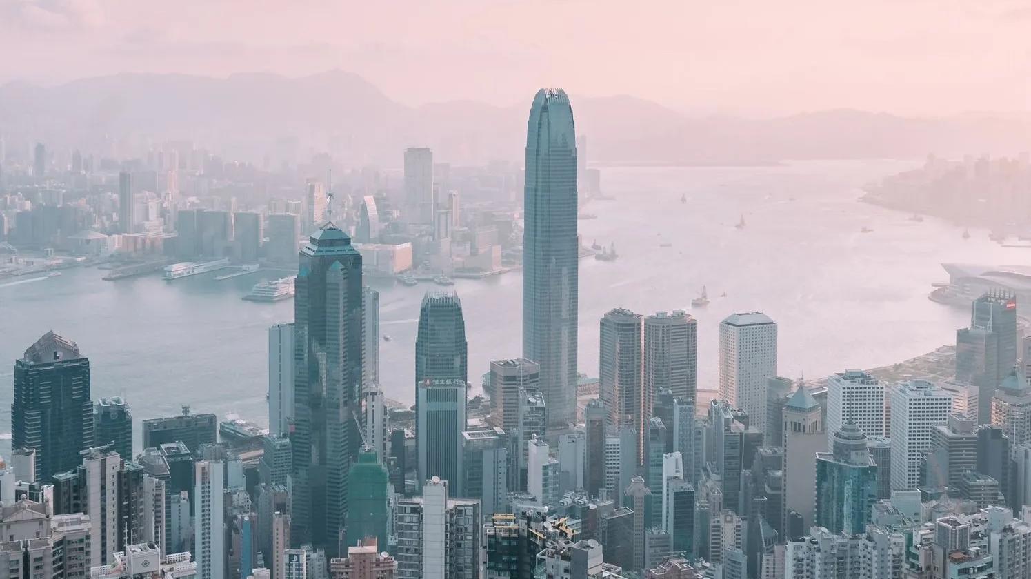 Aerial view of Hong Kong's skyline with tall skyscrapers and Victoria Harbour in the background under a hazy sky.