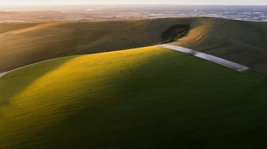 Aerial view of rolling green hills with long shadows cast by the setting sun, creating a serene landscape.