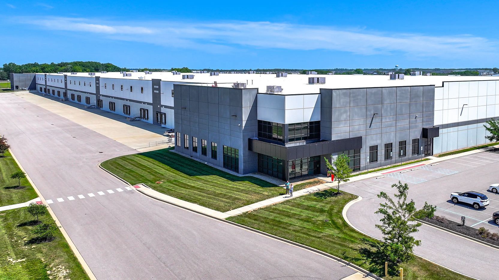 Aerial view of a large modern warehouse or logistics facility with loading docks, parking areas, and surrounding green landscaping.