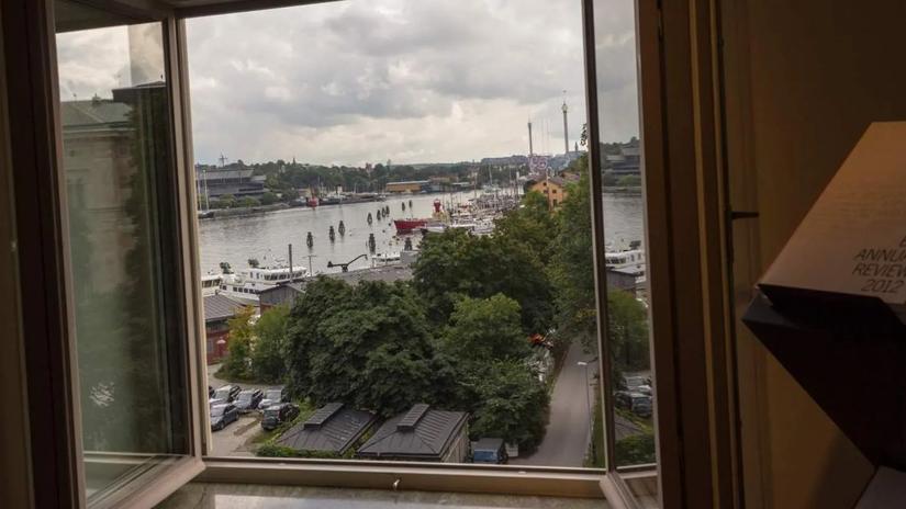 View from an open window overlooking a river with boats, surrounded by trees and buildings under a cloudy sky.