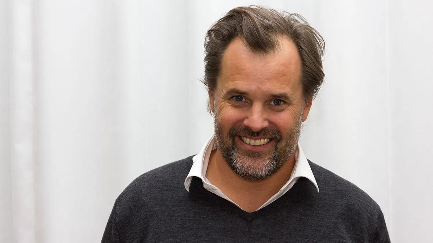 A person with a beard and a friendly smile, wearing a dark sweater over a white collared shirt, standing in front of a light-colored curtain.