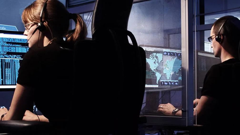 Two people wearing headsets working at computer stations with multiple screens displaying data and a world map.