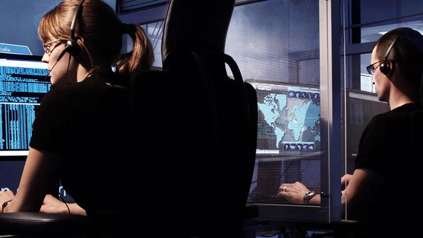 Two people wearing headsets working at computer stations with multiple screens displaying data and a world map.