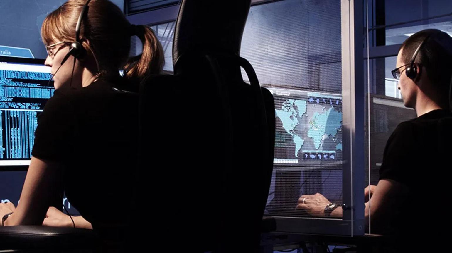Two people wearing headsets working at computer stations with multiple screens displaying data and a world map.