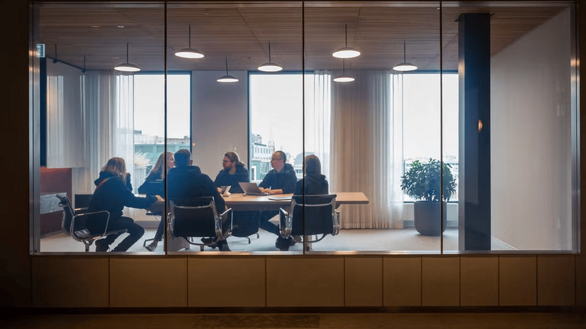 A group of people sitting around a conference table in a modern meeting room with large windows and overhead lights.
