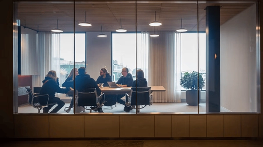 A group of people sitting around a conference table in a modern meeting room with large windows and overhead lights.