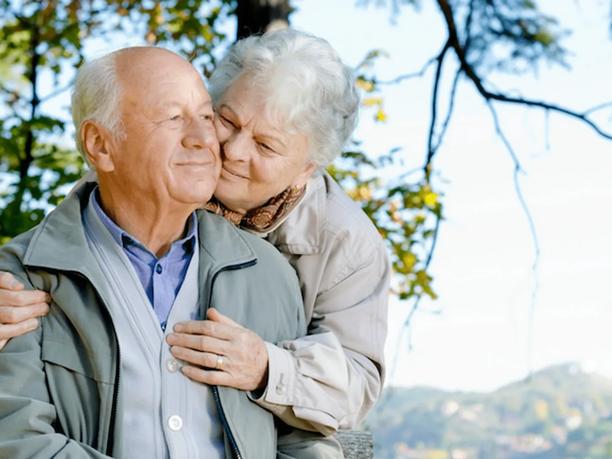 An elderly couple outdoors, with the woman affectionately embracing the man from behind.