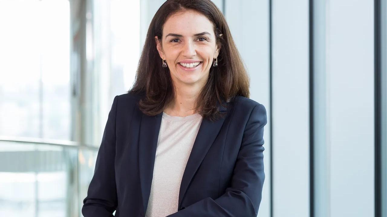 A woman in a dark blazer smiling, standing in a modern office setting with large windows.