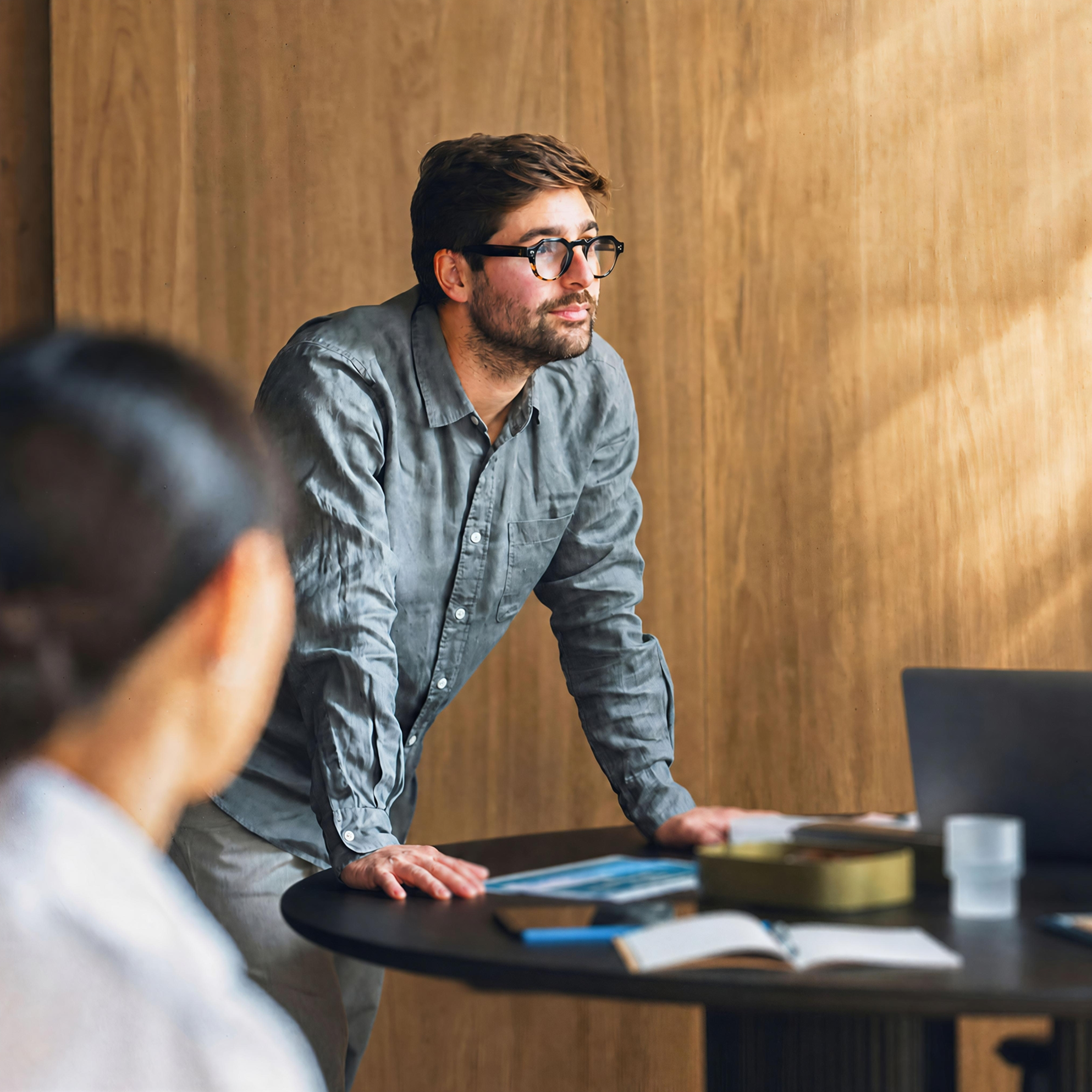 A man with glasses and a beard standing at a round meeting table, leaning forward on his hands while looking toward the side during a discussion.
