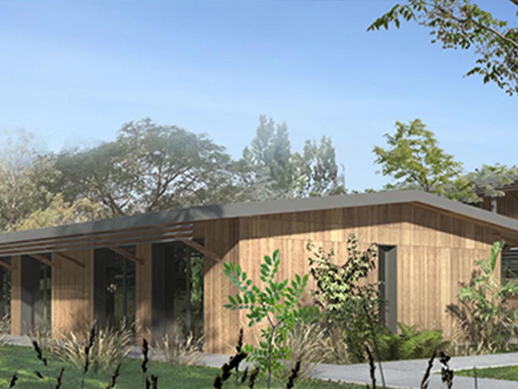 A modern wooden building with a sloped roof, surrounded by lush greenery and trees under a clear blue sky.