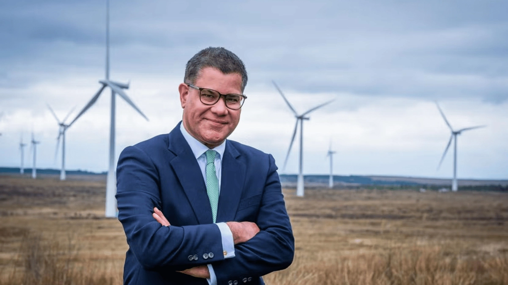 Sir Alok Sharma standing confidently in front of a field of wind turbines under a cloudy sky.