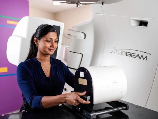 A woman in a blue shirt operates a medical device labeled "TrueBeam" in a clinical setting.
