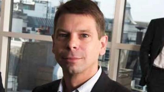 A man in a suit with short hair, standing in an office setting with large windows in the background.