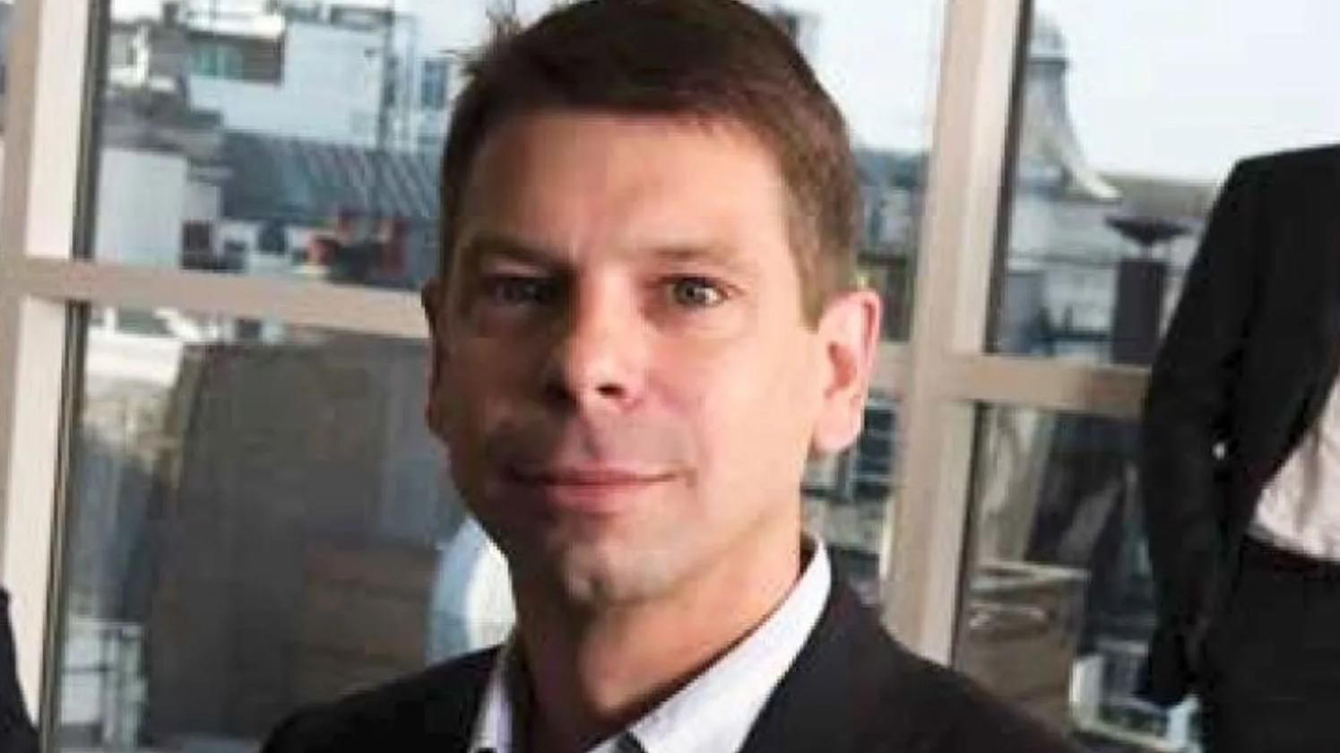 A man in a suit with short hair, standing in an office setting with large windows in the background.