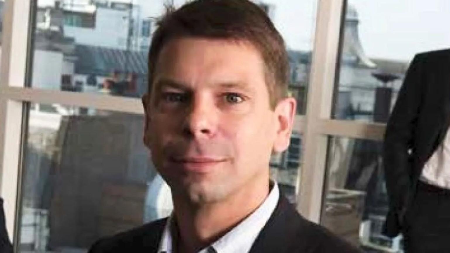 A man in a suit with short hair, standing in an office setting with large windows in the background.