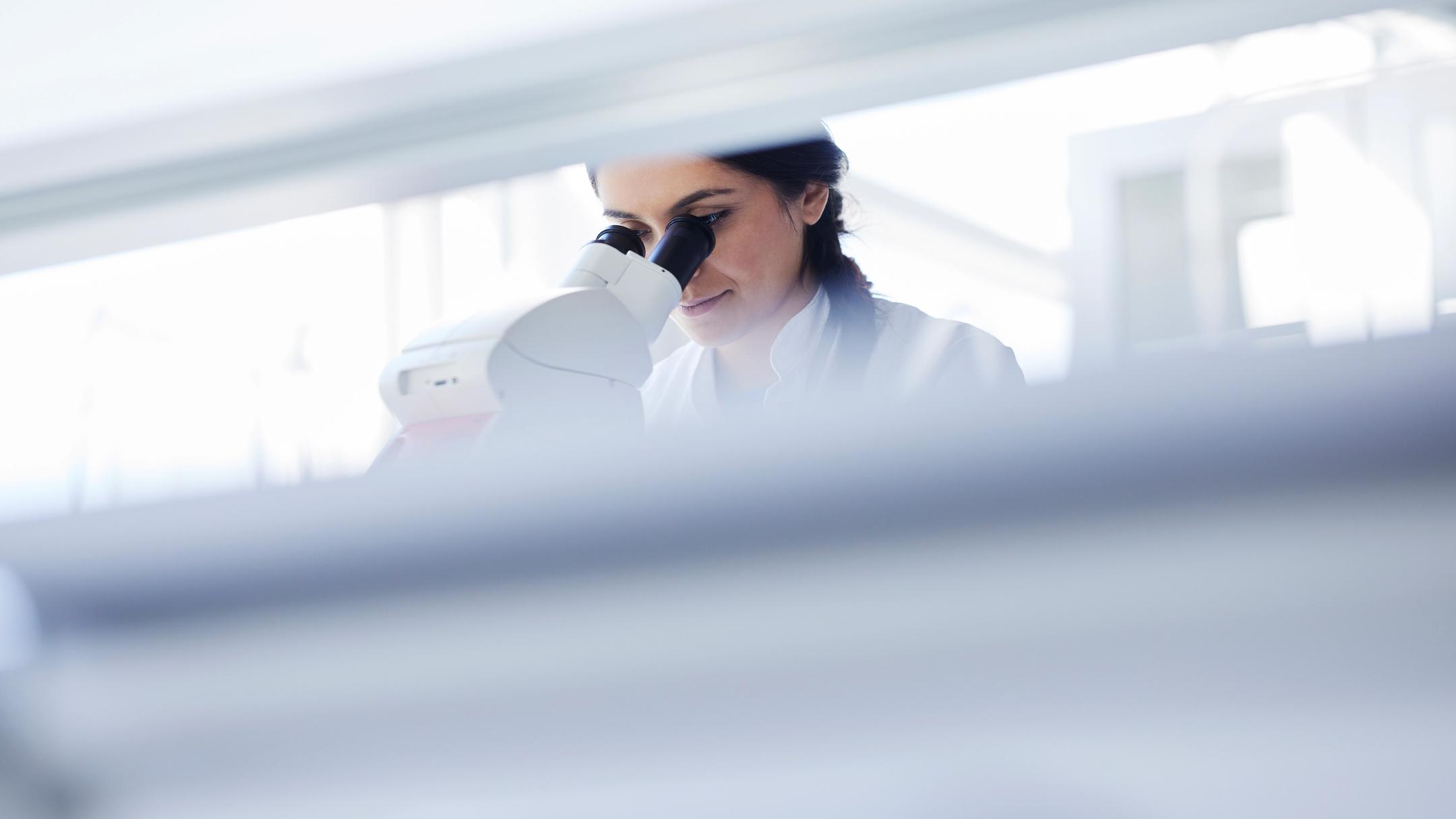 A female scientist looking into a microscope in a brightly lit laboratory.